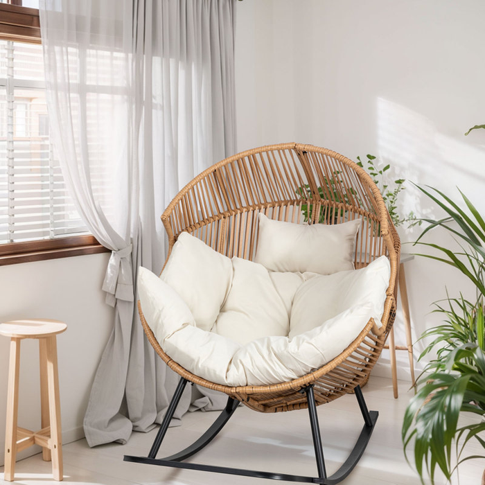 A rattan egg chair with plush white cushions, set in a cozy room filled with natural light. The space is adorned with plants, soft sheer curtains, and a wooden stool, creating a relaxed atmosphere.