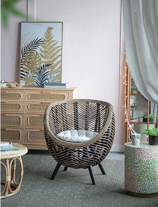 A room featuring a wicker chair, a comfy cushion, a wooden dresser, a plant, and a framed plant print hanging on the wall, creating a cozy atmosphere.