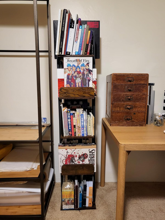 A narrow bookshelf packed with a variety of books and manga, sitting next to a wooden desk with a small storage cabinet