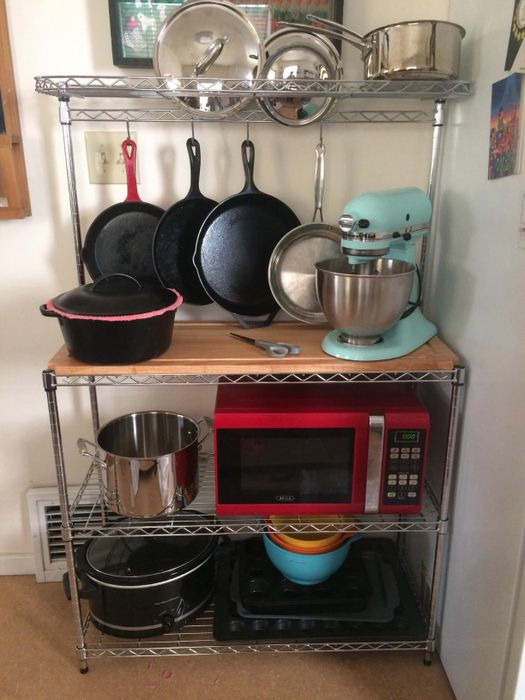 A kitchen shelf holding various cookware, such as cast iron pans, a stand mixer, pots, and a red microwave.