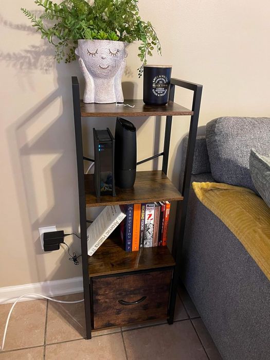 A small wooden shelf beside a gray couch, featuring a potted plant, books, a black speaker, a mug, and a drawer at the base