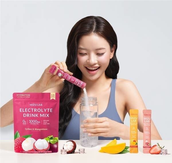 A woman smiling in a tank top is pouring Mizu Lab's electrolyte drink mix into a bottle, with flavors like lychee & mangosteen and orange visible nearby