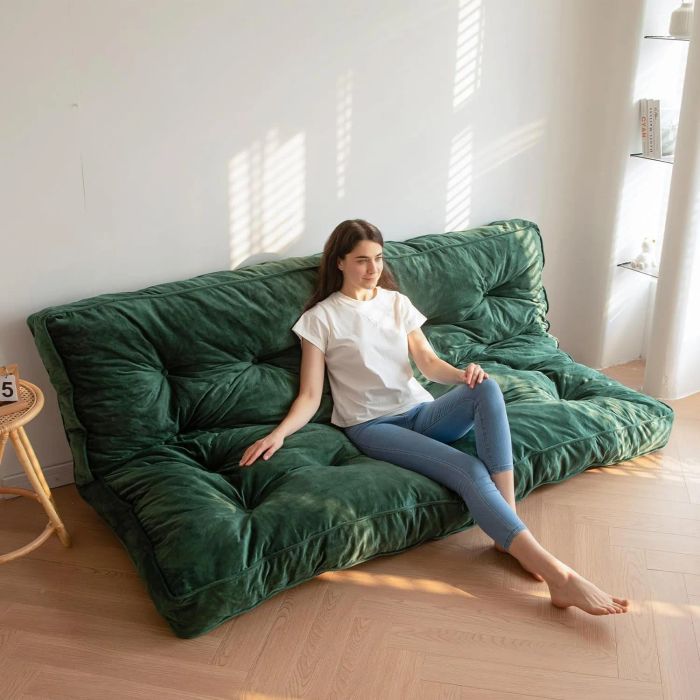 A woman lounging on a green, plush, tufted sofa in a minimalist living room, dressed in a white t-shirt and jeans, with her bare feet resting on the floor.