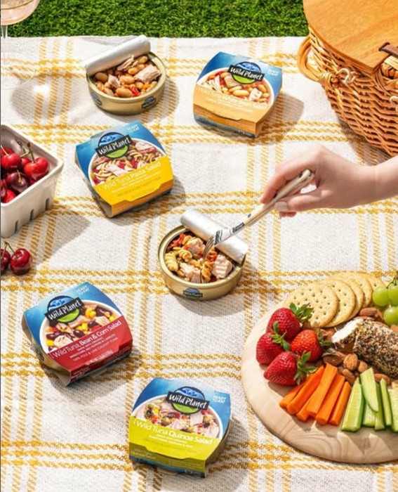 A picnic scene with a hand opening a can of Wild Planet tuna on a checkered blanket, surrounded by fresh fruit, crackers, and vegetables