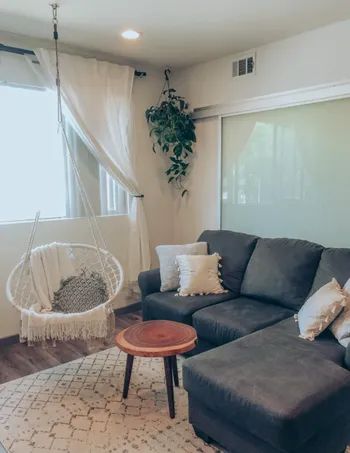 A cozy living room featuring a gray sectional sofa, a round wooden coffee table, a hanging chair, throw pillows, and a plant by the window. No people are in the image.
