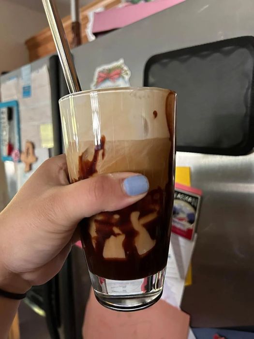 A hand holds a glass of swirled chocolate iced coffee with a straw, positioned in front of a refrigerator.