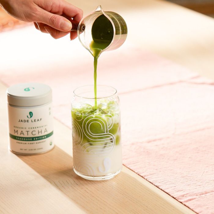 A hand pouring matcha into a glass of milk next to a container of Jade Leaf matcha, placed on a table
