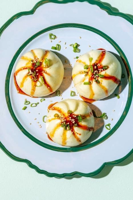 Three round bao buns on a white plate with green trim, garnished with green onions, sesame seeds, and drizzled with sauce