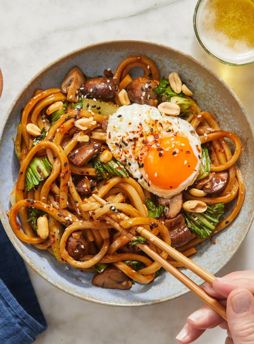 A close-up of a bowl of stir-fried noodles with vegetables, mushrooms, peanuts, and a fried egg on top. A hand is seen picking up the noodles with chopsticks.