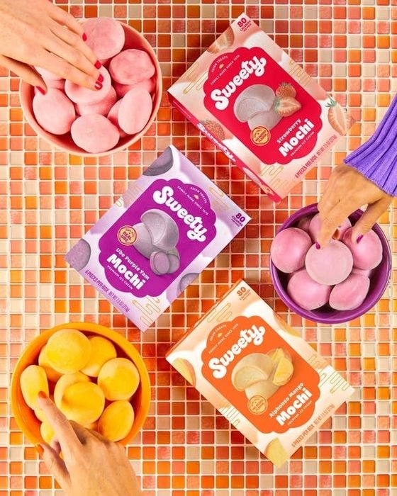 A person's hand reaching for different flavors of packaged mochi on a tiled surface. Boxes include 