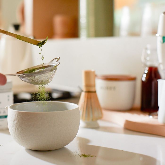 A person sifting matcha powder into a bowl on a kitchen counter, with tea accessories and a container in view