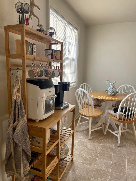 A kitchen corner featuring a wooden shelf filled with appliances, hanging cups, and a table with four chairs topped with a floral teapot.
