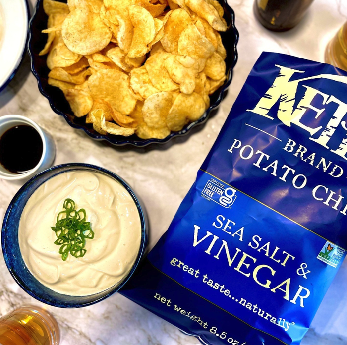 A bag of Kettle Brand sea salt and vinegar potato chips placed on a table next to dip and other snacks