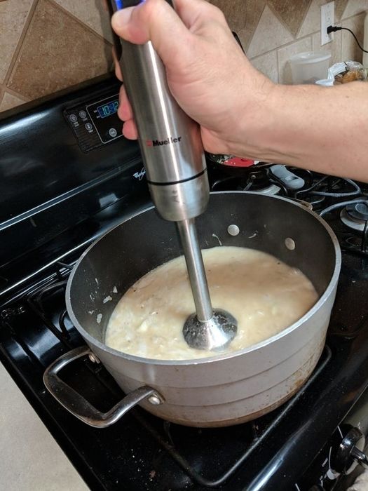 A person's hand using a Mueller immersion blender to blend soup in a large pot on a stove