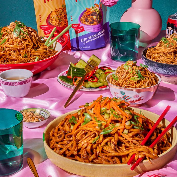 A variety of noodle dishes styled with Milo instant noodle packs, topped with vegetables, and surrounded by drinks and condiments on a table