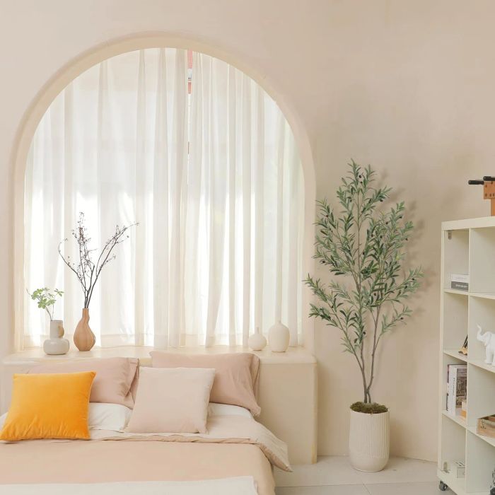 A minimalist bedroom featuring a bed with light-colored pillows and sheets, an arched window adorned with sheer curtains, plants, and a white bookshelf displaying decor items