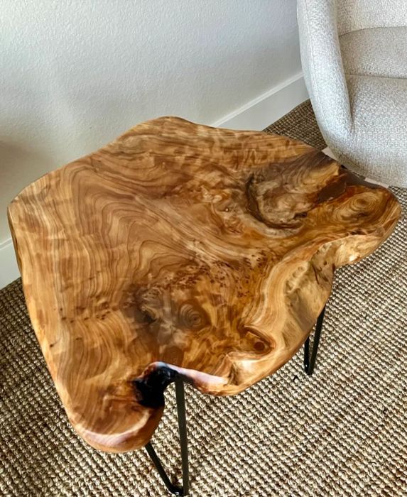 A natural wood accent table with a unique, irregular shape sitting beside a modern, upholstered chair. The table features beautifully intricate wood grain patterns.