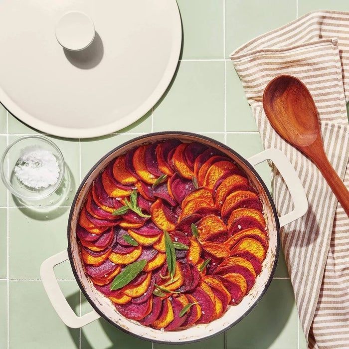 A round casserole filled with layers of sliced vegetables, topped with aromatic herbs, next to a wooden spoon and a striped napkin.