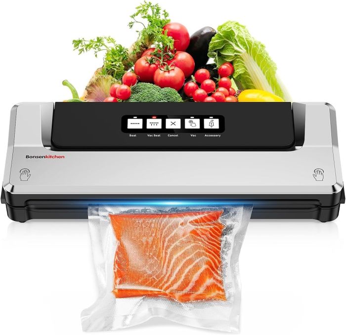 A Bonsenkitchen vacuum sealer in action, sealing a bag filled with sliced meat. Fresh vegetables like tomatoes, lettuce, and broccoli are placed behind the machine