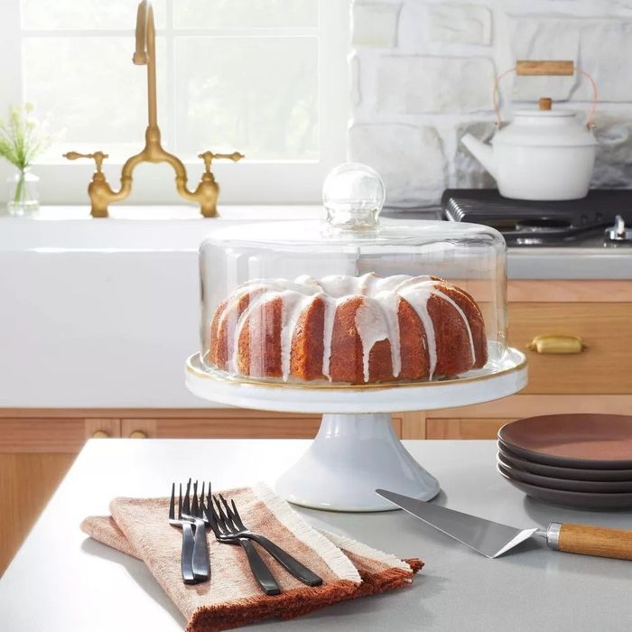 A Bundt cake, topped with icing, sitting on a white cake stand under a glass dome in a kitchen setting, surrounded by plates and utensils.