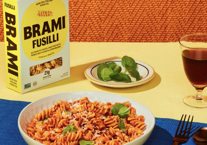 Brami fusilli pasta box placed beside a plate of cooked fusilli, topped with basil and cheese, accompanied by a drink and more basil on a table