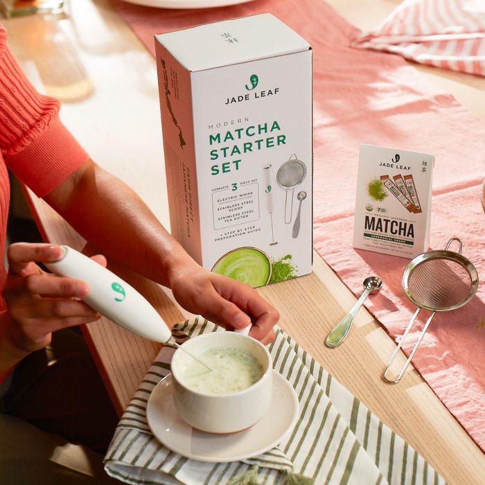 A person using a milk frother to prepare a matcha latte, with the Jade Leaf Matcha Starter Set and ingredients laid out on the table