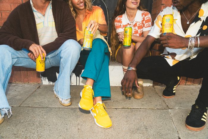 Four people are enjoying a sunny day on a sidewalk, each holding a can of Guayakí Yerba Mate, dressed casually in summer clothes and sneakers