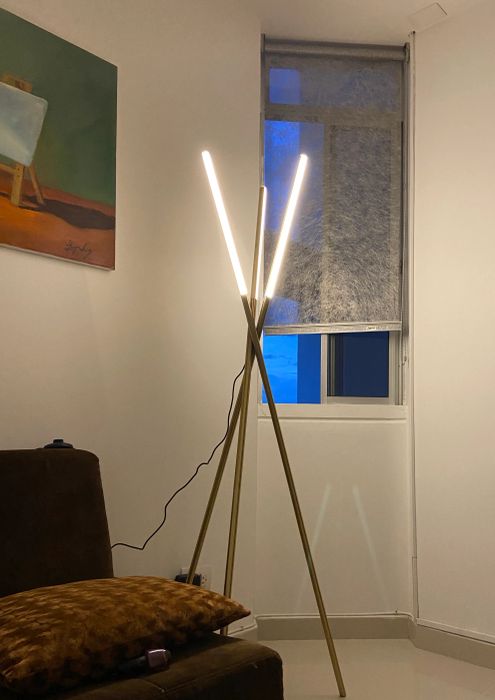 A modern living room featuring a tripod floor lamp, a wall painting, a partially closed blind on a window, and a TV on a stand.