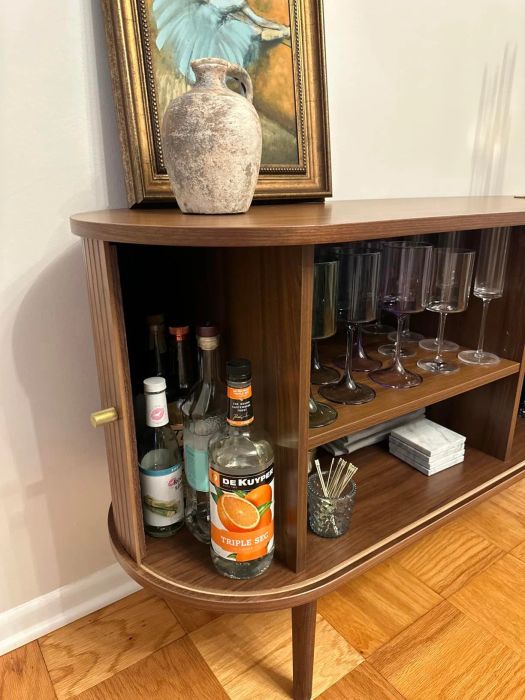 A wooden cabinet showcasing a collection of glassware and bottles, including De Kuyper Triple Sec, with a rustic vase and a framed painting placed on top.