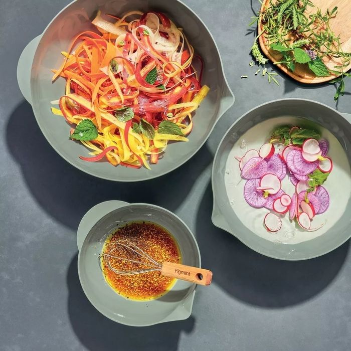 Three bowls on a gray surface: one filled with spiralized vegetables, another with radishes in water, and the last with a freshly whisked dressing. A wooden board holds fresh herbs and greens nearby.
