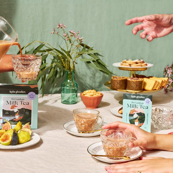 Milk tea being poured into glass cups on a table with snacks and pink flowers in the background. Packages of milk tea are visible