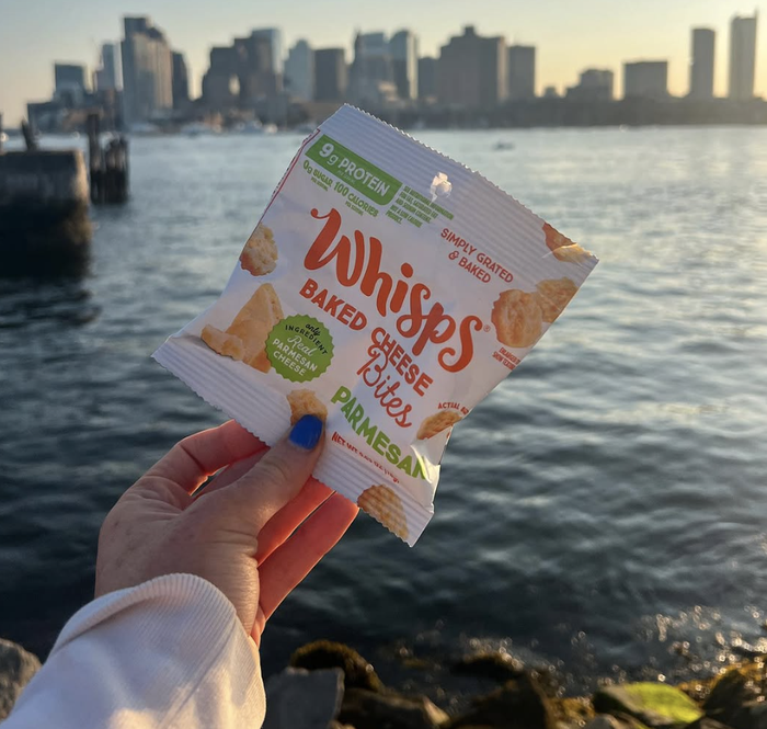 A person holds a bag of Whisps Baked Cheese Bites, with a city skyline and water in the background