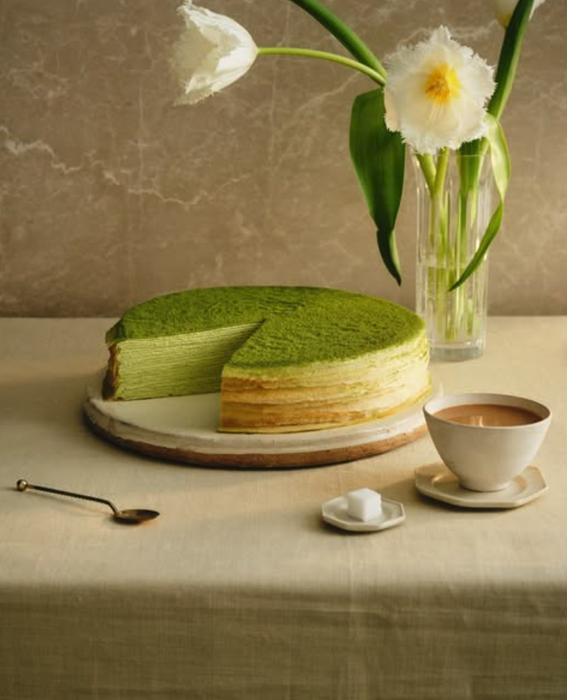 A slice of Matcha Mille Crepe cake missing from the whole, resting on a table with a cup of tea, sugar cubes, and a vase with flowers nearby