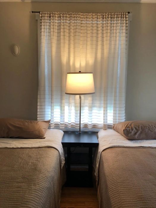 A bedroom setup with two single beds placed next to each other, with a nightstand and lamp between them, complemented by a window and curtain in the background.