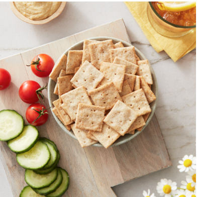 A wooden board is beautifully arranged with a bowl of square crackers, surrounded by cherry tomatoes, cucumber slices, a bowl of dip, and a drink on the side