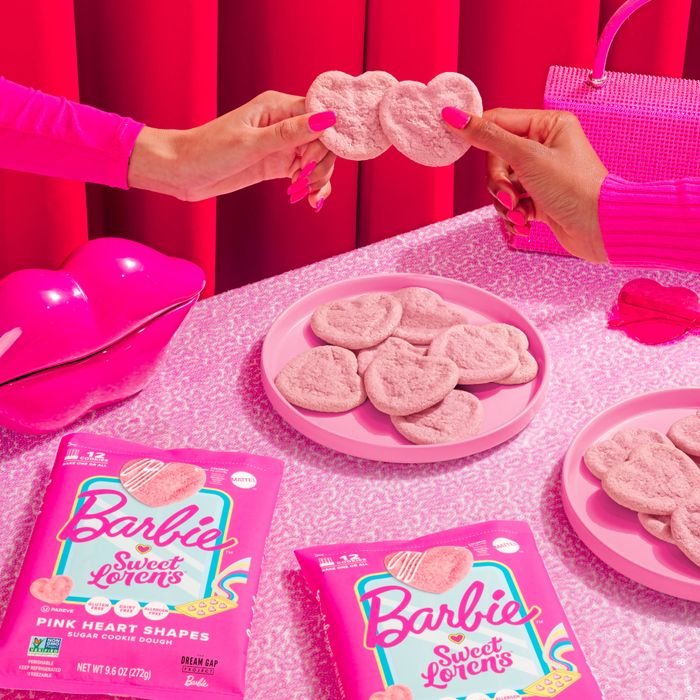 Two hands hold a heart-shaped pink cookie above a table decorated with Barbie-themed cookie packaging and accessories, evoking a playful shopping vibe