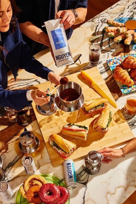 A person scoops Nguyen Coffee Supply coffee grounds into a pot, surrounded by sandwiches, croissants, donuts, and cans of Nguyen Coffee Supply coffee on a table