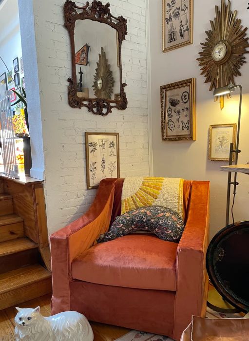 A vintage-inspired corner of the living room with an orange velvet armchair, a decorative mirror above, wall art, and a white cat statue.