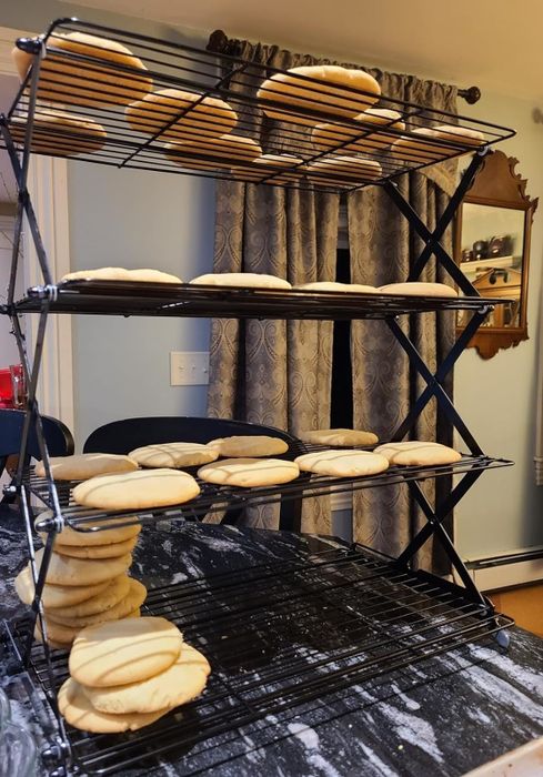 Freshly baked cookies cooling on stacked wire racks in a kitchen, with a pile of cookies sitting on the countertop