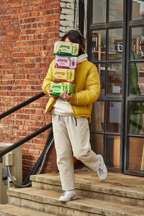 A person carrying four boxes of Sanzo drinks as they descend a set of concrete stairs outside a brick building, dressed in a yellow puffer jacket and beige pants