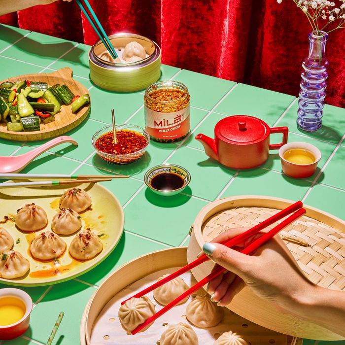 A variety of dumplings and sauces laid out on a dining table, accompanied by steaming pots, a tea set, and a vibrant red backdrop. Hands are seen using chopsticks to serve the dishes