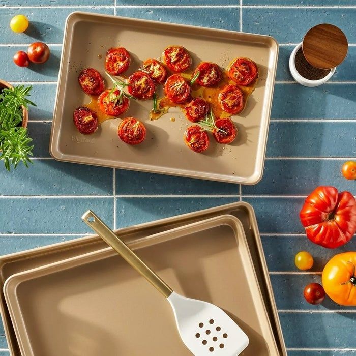 An overhead shot of roasted tomatoes on a baking sheet, with a spatula alongside, surrounded by fresh tomatoes and herbs on a tiled countertop.