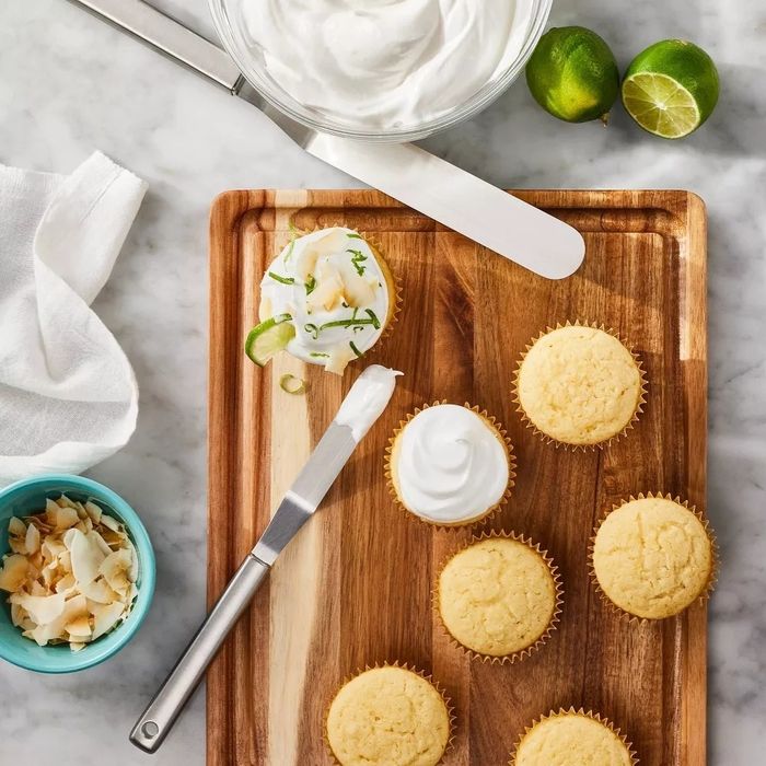 Cupcakes adorned with frosting, a bowl of whipped cream, sliced limes, and toasted coconut chips, all neatly arranged for decoration on a wooden board.