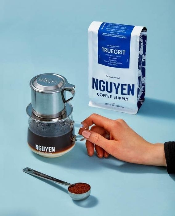 A hand holds a glass of coffee brewed with a stainless steel phin filter. Next to it is a bag of Nguyen Coffee Supply's Truegrit coffee, along with a teaspoon of coffee grounds