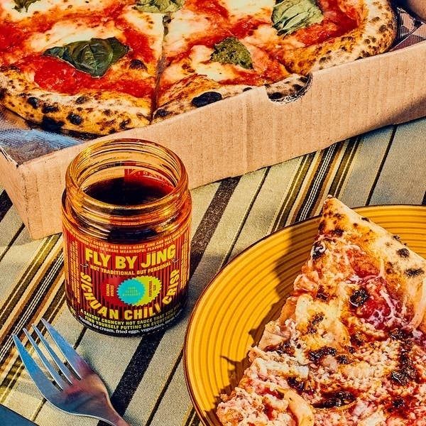 A jar of Fly By Jing Sichuan Chili Crisp sits next to a slice of pizza and a pizza box, with a fork resting nearby on the table