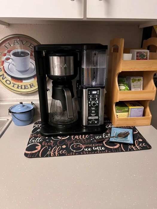 A coffee maker placed on a countertop, accompanied by a sign, tea bags, and a plate of cookies, creating a cozy, well-organized coffee station in the kitchen.