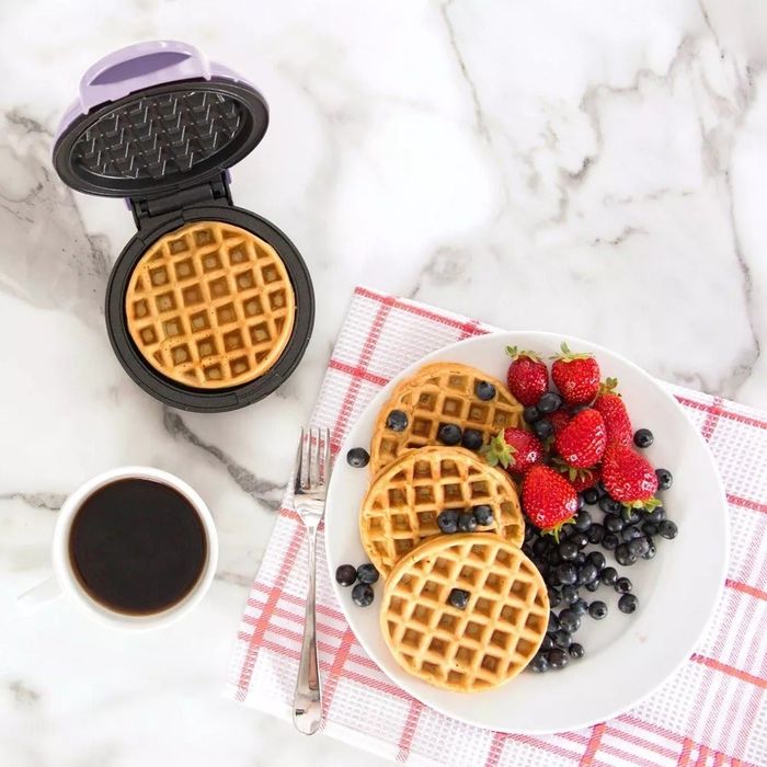 A compact waffle maker with a freshly cooked waffle, accompanied by a plate of waffles, strawberries, blueberries, and a cup of coffee, all placed on a marble surface with a checkered napkin.