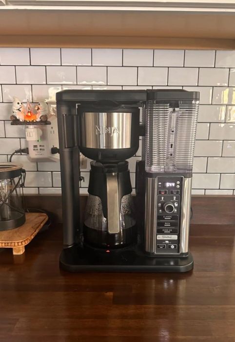 A coffee maker with a digital display sits on a kitchen counter, with a white subway tile backsplash and various coffee accessories in the background.