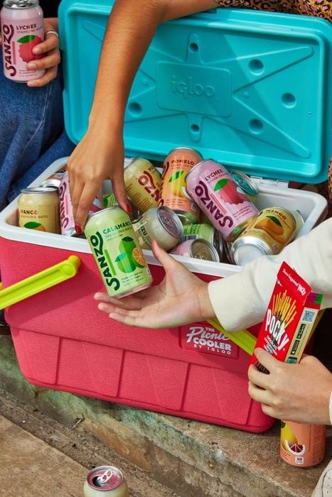 A group of people reaching into a cooler filled with Sanzo sparkling water cans, with one person also holding a pack of Pocky sticks
