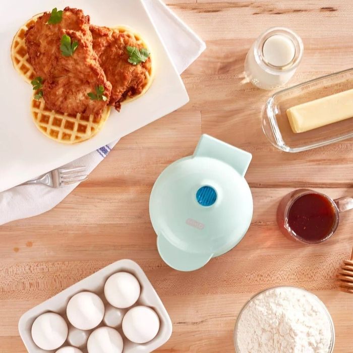 A plate of chicken and waffles, with a milk bottle, butter dish, syrup, eggs, flour, and a mini waffle maker arranged on a wooden table.
